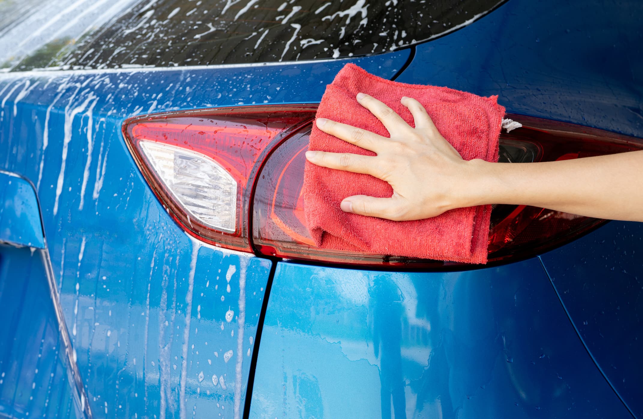 Car wash service. Car covered with white soap foam. Man hand holding red microfiber cloth and polish tail light of blue car. Auto care service business concept. Man cleaning and detailing luxury car.