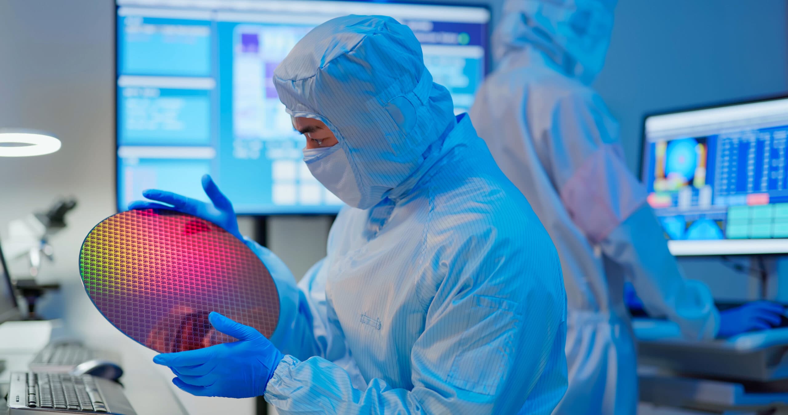 asian male technician in sterile coverall holds wafer that reflects many different colors with gloves and check it at semiconductor manufacturing plant